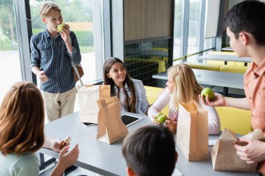 happy teenagers eating sandwiches and apples while talking in school dining room