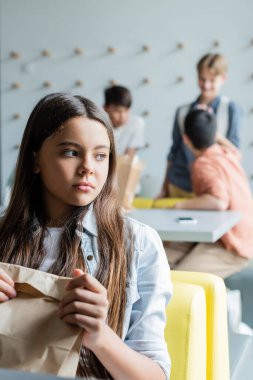 frustrated girl sitting with paper bag in dining room near classmates on blurred background clipart