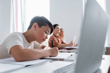asian schoolboy writing in notebook near blurred laptop and classmates clipart