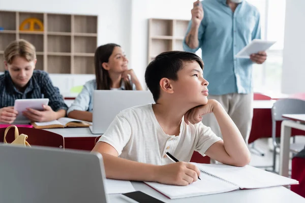 Asian Schoolboy Looking Away While Writing Notebook Laptop Classroom — 스톡 사진