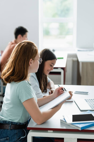 Schoolgirl writing on notebook near gadgets and smiling classmate 