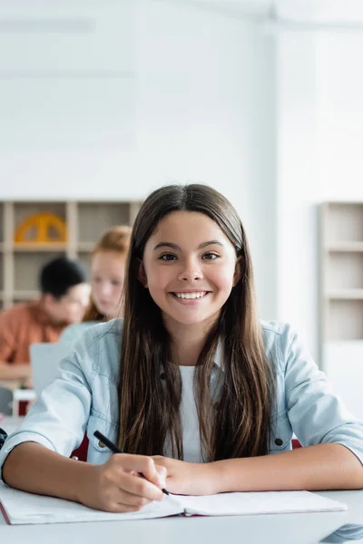 Teenager with pen smiling at camera near notebook in classroom