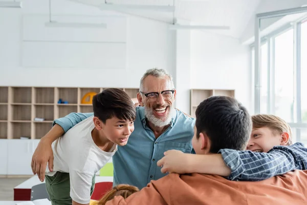 Cheerful Multiethnic Boys Embracing Laughing Middle Aged Teacher Classroom — Stock Photo, Image