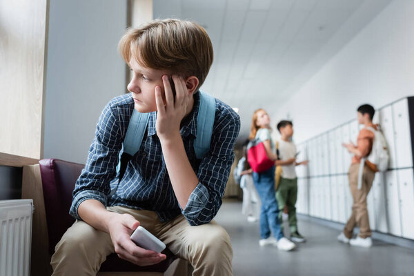 depressed boy with smartphone sitting alone in school corridor near teenagers on blurred background