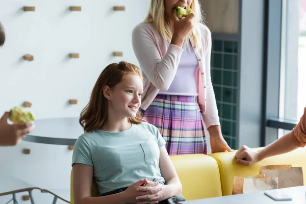 学校でリンゴを食べる友達の近くで笑顔の赤毛の女子高生 — ストック写真