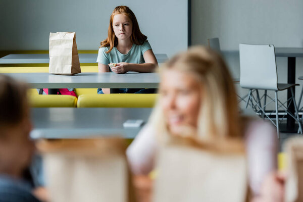 depressed girl with sandwich sitting alone in school eatery near classmates on blurred foreground