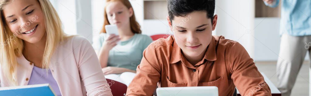 Schoolkid looking at blurred digital tablet near smiling classmate, banner 