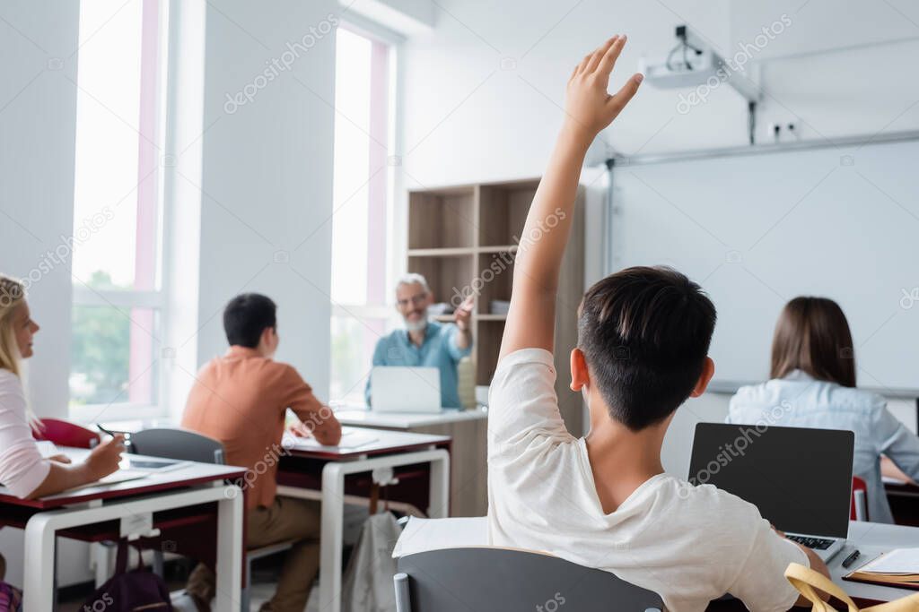 Pupil raising hand near laptop and blurred classmates 