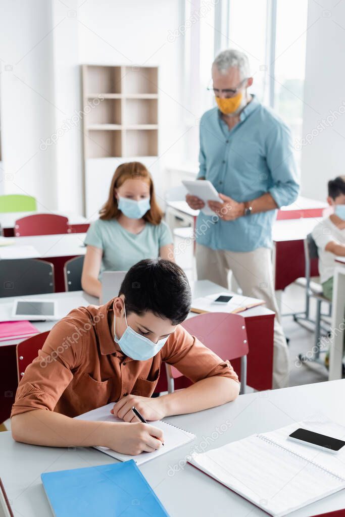 Schoolkid in medical mask writing on notebook in classroom 