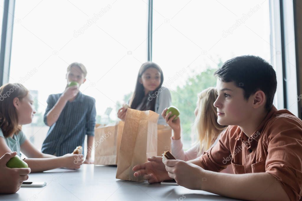 schoolboy holding sandwich during lunch break in dining room near classmates