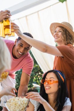 cheerful multiethnic friends clinking bottles of beer during party in patio clipart