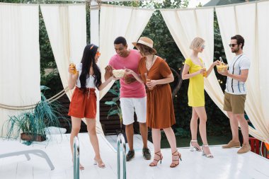 Çok ırklı arkadaşlar havuz başında bira içiyor ve abur cubur yiyorlar.