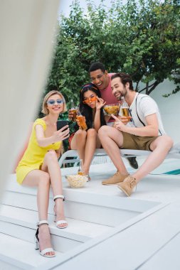 Partide çok ırklı arkadaşlarıyla cep telefonundan selfie çeken mutlu kadın, bulanık önplan