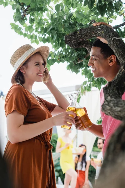 stock image african american man and smiling woman in straw hat clinking bottles of beer near blurred friends