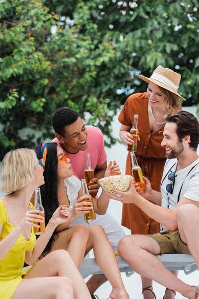 Улыбающийся мужчина держит миску попкорна рядом с веселыми межрасовыми друзьями, пьющими пиво