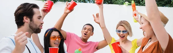 在聚会期间 快乐的男人在多文化的朋友面前展示出了胜利的姿态 他拿着塑料杯和横幅敬酒 — 图库照片
