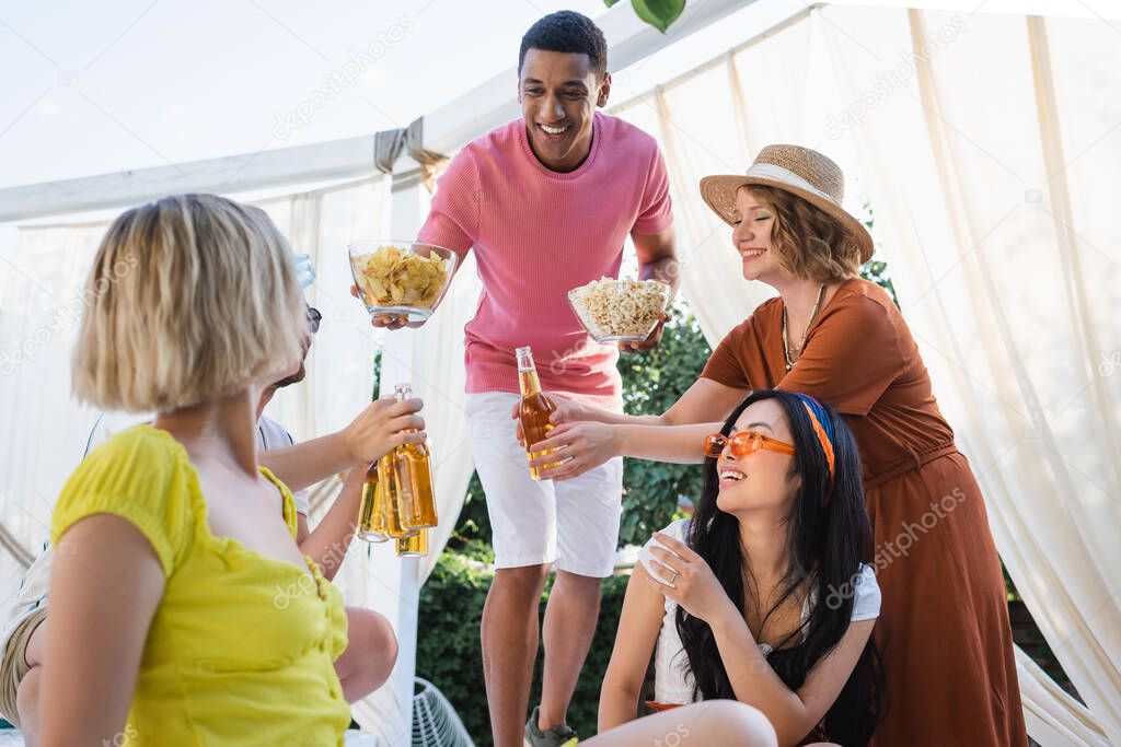 happy african american man proposing chips and popcorn to multiethnic friends drinking beer in patio