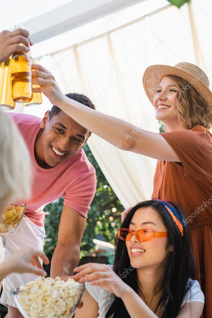 cheerful multiethnic friends clinking bottles of beer during party in patio