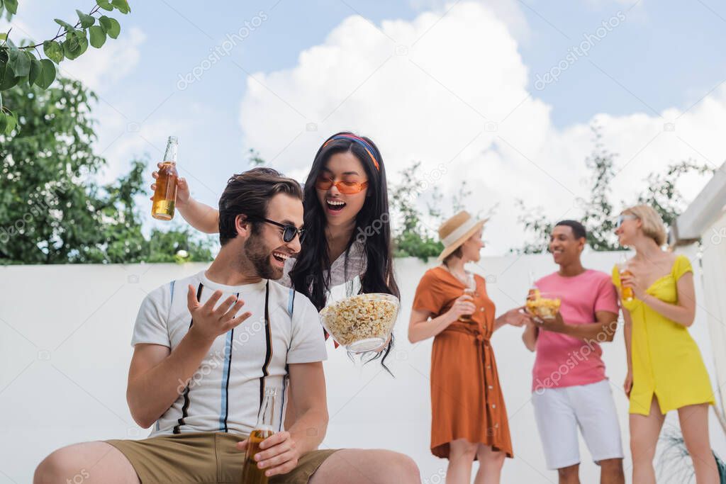 asian woman proposing popcorn to man near multiethnic friends drinking beer on blurred background