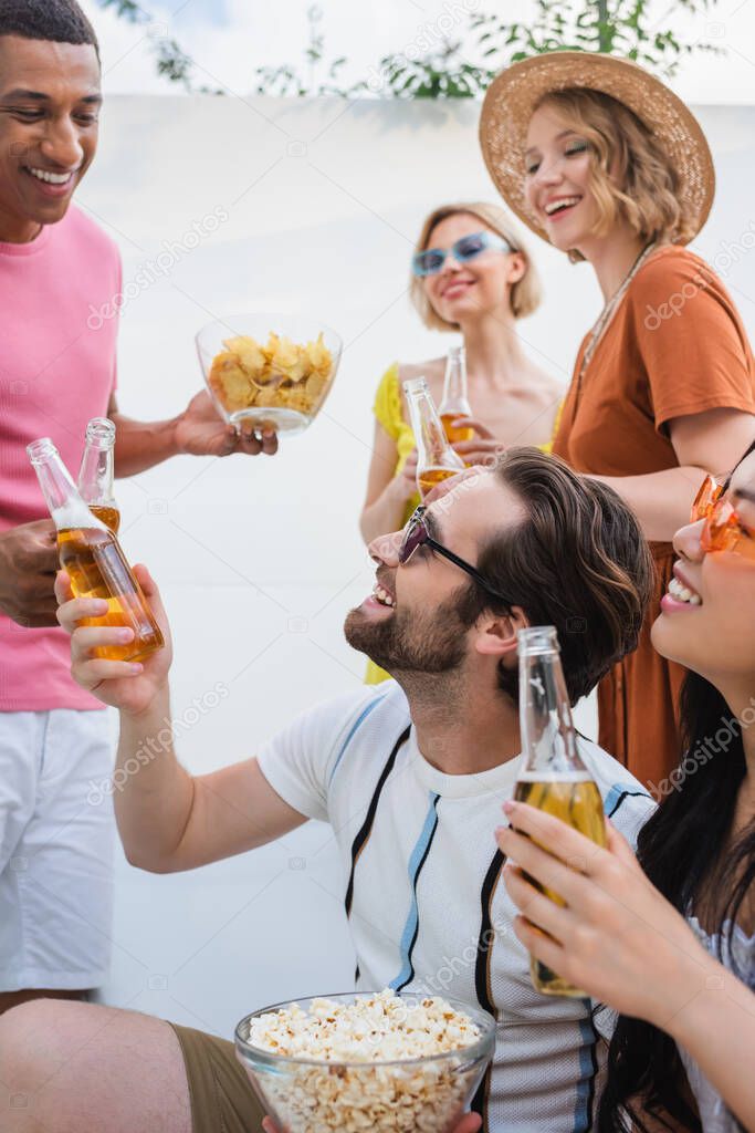 happy interracial men clinking with bottles of beer near smiling women 