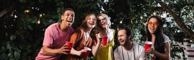 excited multicultural friends singing karaoke during party in night garden, banner clipart