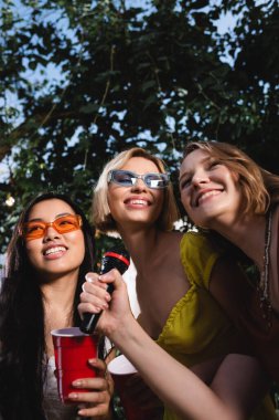 Low angle view of smiling multiethnic women with plastic cup holding microphone outdoors during party  clipart