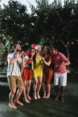 Genç çok ırklı arkadaşlar karaoke yapıyor ve akşamları çimlerin üzerinde plastik bardaklar tutuyorlar. 