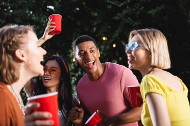 Excited african american man showing yes gesture and holding plastic cup near friends during party  clipart