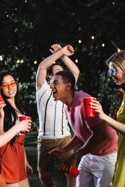 Pozitif Afro-Amerikalı adam yaz partisinde ırklar arası arkadaşlarıyla plastik bardak tutarak dans ediyor. 
