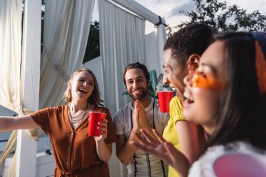 Positive interracial people with plastic cups having fun near patio clipart
