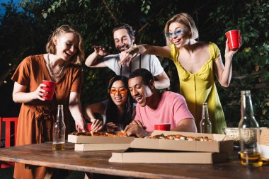 Açık havada pizzanın yanında oturan plastik bardaklı mutlu ırklar arası arkadaşlar. 