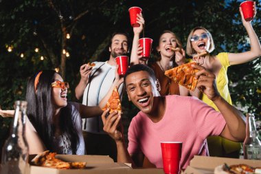 Gülümseyen Afrikalı Amerikalı adam dışarıda plastik bardakları olan bulanık arkadaşlarının yanında pizza tutuyor. 