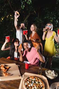 Yaz partisinde elinde pizza ve plastik bardaklar olan çok ırklı mutlu arkadaşlar. 