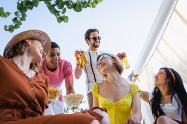 Verandada bira ve yiyeceklerle arkadaşlarının yanında oturan, gülümseyen, etnik çeşitliliğe sahip kadınlara bakış açısı düşük. 