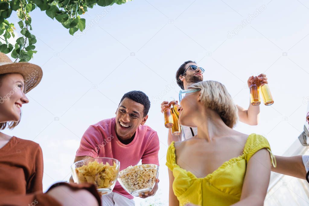 Low angle view of cheerful african american man holding snacks near friends outdoors 