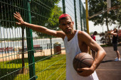 Afroamerikanischer Sportler mit gefärbten Haaren hält Basketballball am Zaun auf dem Platz 