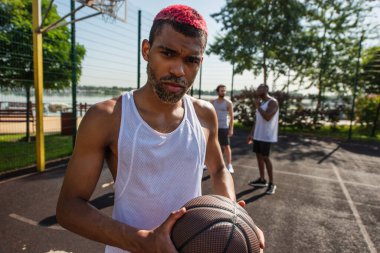 African american basketball player holding ball near blurred men on playground  clipart