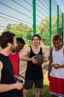 Açık havada akıllı telefonları olan ırklar arası arkadaşlarının yanında basketbol topu tutan gülümseyen bir adam. 