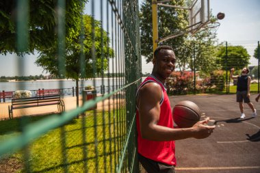 Young african american sportsman holding basketball ball near blurred fence on playground  clipart