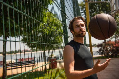 Young man looking at camera while throwing basketball ball on court  clipart