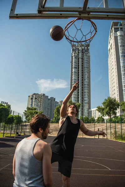 友人の近くにジャンプ若いスポーツマン 遊び場のバスケットボールのフープとボール — ストック写真