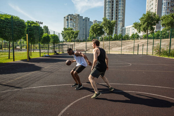 Side view of african american man with basketball ball running near friend on playground 