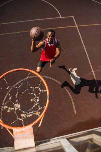 興奮したアフリカ系アメリカ人スポーツマンの高い角度ビュージャンプとともにバスケットボールボール近くフープ — ストック写真