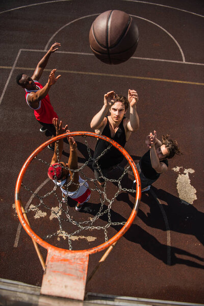 Overhead view of man throwing basketball ball near multiethnic friends with raised hands and hoop 