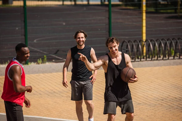 Glimlachende Sportman Met Basketbal Wijst Met Vinger Naar Afrikaanse Amerikaanse — Stockfoto