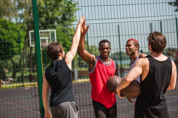 Uśmiechnięci Sportowcy Międzyrasowi Piłką Koszykówki Przybijają Piątkę Pobliżu Ogrodzenia — Zdjęcie stockowe