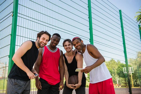 Веселые Многонациональные Спортсмены Баскетбольным Мячом Смартфонами Смотрящие Камеру Улице — стоковое фото