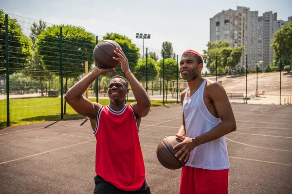 Gülümseyen Afro-Amerikan sporcusu oyun alanında arkadaşının yanında basketbol topu tutuyor. 