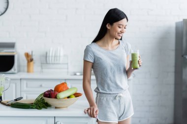 happy asian woman standing with glass of homemade smoothie near bowl with fresh vegetables and apples clipart