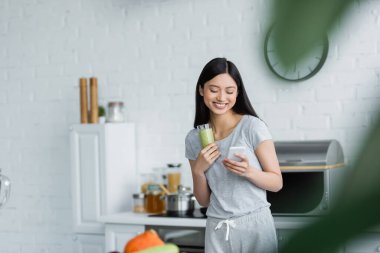 Neşeli Asyalı kadın bir bardak smoothie ile cep telefonuyla ön planda bulanık bir şekilde mesajlaşıyor.